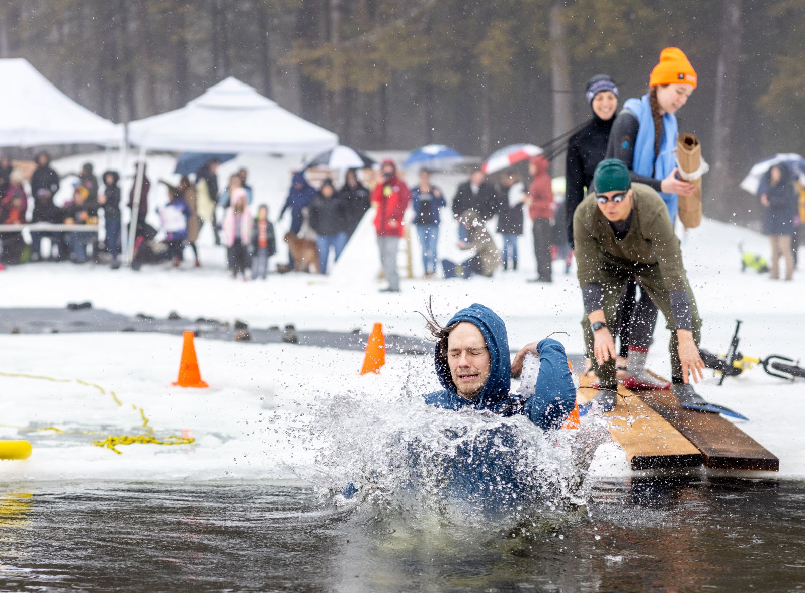 A man in a blue hooded sweatshirt grimaces from the cold water while others line up behind him to plunge; a large crowd looks on in the background