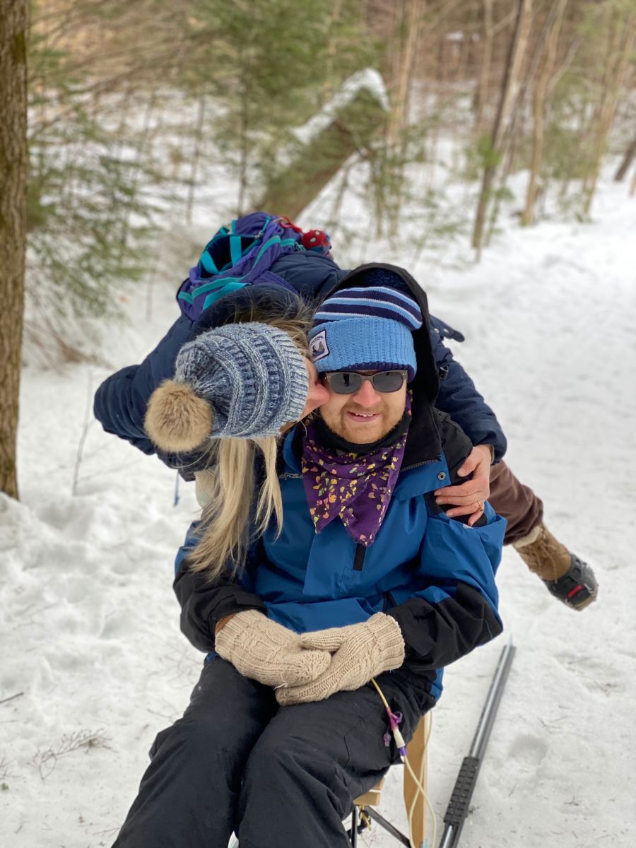 A young man on a kicksled smiles as his mother leans over to kiss his cheek